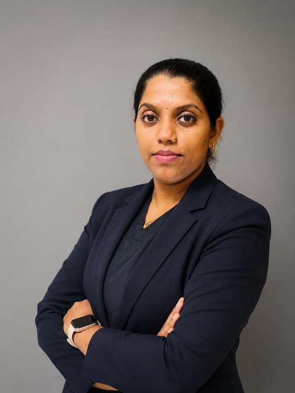 Asha Rani PS in a navy blazer, arms folded, against a plain studio backdrop